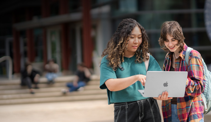 Two university students looking at a MacBook screen while standing on campus
