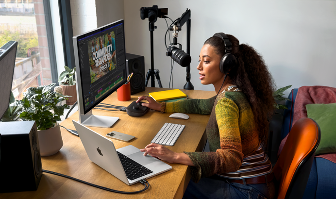 Μια γυναίκα στο σπίτι της δουλεύει στο γραφείο της με το MacBook Pro συνδεδεμένο σε δύο εξωτερικές οθόνες, φορά ακουστικά και μιλά στο μικρόφωνο