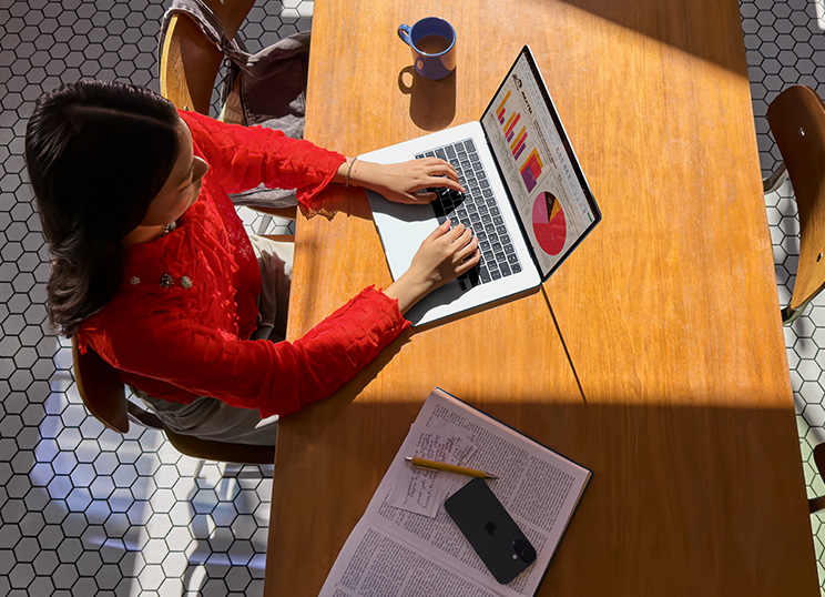Seseorang menggunakan MacBook di meja, dengan cangkir, ponsel pintar, dan kertas di dekatnya.