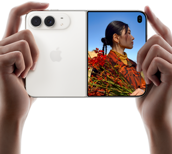 iPhone Duo, zvaigžņu baltā krāsā, tiek turēts divās rokās horizontālā novietojumā, atvērts, kreisajā panelī redzamas 48 MP Dual Fusion aizmugurējās kameras, labajā panelī redzams, kā ārējā displejā tiek uzņemta cilvēka bilde, fotografējamais redz savu seju, kamēr tiek uzņemta bilde ar Duo Preview
