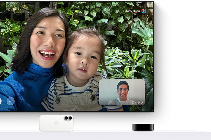 جهاز Apple TV 4K وجهاز iPhone: شاشة تلفزيون تعرض مكالمة فيس تايم، شخص يحمل طفلاً صغيراً، المتصل الثاني يظهر في إطار