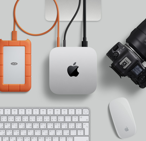 لقطة علوية لجهاز Mac mini فضي اللون، مع سلكين متصلين من المنافذ الخلفية بشاشة، وبجواره ماجيك ماوس أسود ولوحة مفاتيح ماجيك سوداء