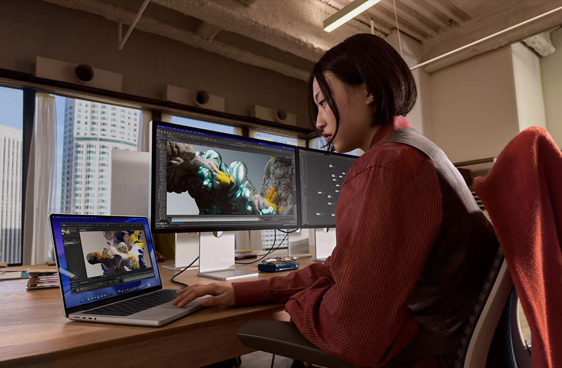 A person works at a desk with a MacBook Pro connected to external display, both screens showing a 3D render, buildings visible through the window behind