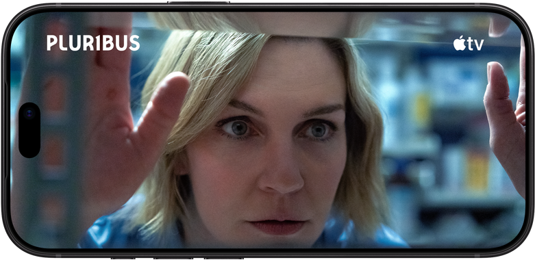iPhone 18 Pro na šírku zobrazuje detailný záber ženy, ktorá si prezerá policu. Názov seriálu: Pluribus