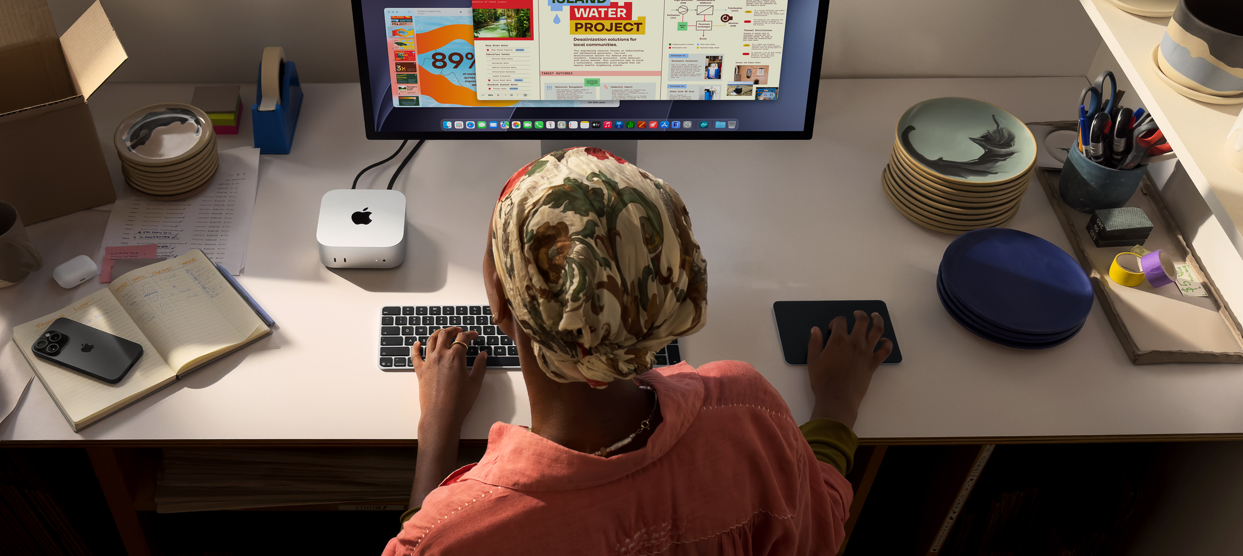 Masada oturan bir kişi Mac mini ile yazı yazıyor, Studio Display bağlı, Magic Keyboard, Magic Trackpad ve çeşitli eşyalar