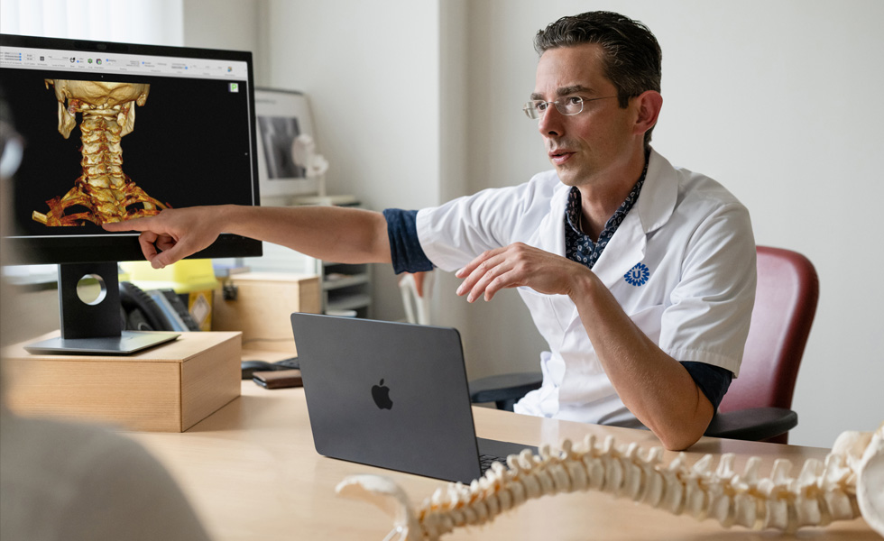 Een medisch professional wijst naar een 3D-skeletweergave van de achterkant van een nek op een Studio Display, terwijl een MacBook op het bureau staat